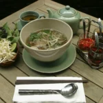 Pho with herbs, chili and a pot of tea on a wooden table