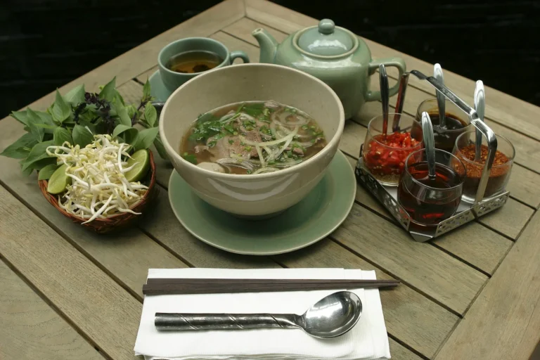 Pho with herbs, chili and a pot of tea on a wooden table